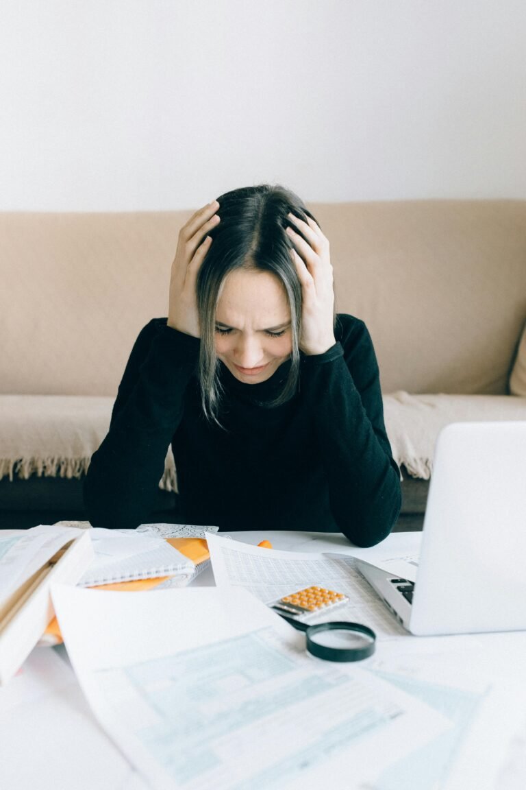 A person holding their head in their hands over a desk covered in tax documents and a laptop